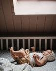 A baby in a white bonnet and light clothes lies wrapped in the mushie Organic Cotton Muslin Swaddle Blanket on a patterned bed, bathed in soft natural light from above.