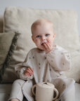 A baby sits on a beige couch, wearing a mushie Long Sleeve Bib and a light-colored outfit with tiny prints, holding food and eating, with a small cup in front. A green pillow is visible in the background.