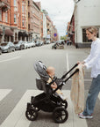 A woman pushes a baby wrapped in a mushie Organic Cotton Muslin Swaddle Blanket across a busy city crosswalk, while the baby gazes up at her amid parked cars and buildings.