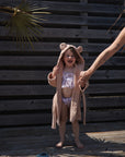A child in a mushie Bear Robe made of organic cotton, featuring a hood and ears, stands on a wooden deck holding an adult's hand. The child wears a floral swimsuit as palm leaf shadows fall across them in the sunlight.