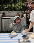 A young child wears the mushie Bear Robe while eating at an outdoor table set with pastel dishes. A woman in a white dress stands nearby, with a striped tablecloth on the table and blooming flowers in the background.