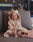 A young child in a mushie Bear Robe made of beige organic cotton with cute ears smiles on a gray couch, holding a toy. A window behind reveals blurred figures and an inviting outdoor view.