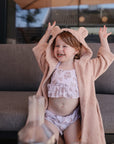 A young child in the mushie Bear Robe sits on a gray outdoor couch, smiling with arms raised happily over a floral swimsuit.