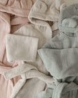 Three mushie Bear Robes in pastel pink, beige, and light sage green organic cotton are side by side. Each robe has cute bear ears on the hood, with a matching washcloth atop the beige Bear Robe.