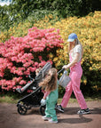 A woman in pink overalls pushes a stroller beside a girl in green, walking past flowering bushes, with a mushie Water Resistant Wet Bag visible in their diaper bag.