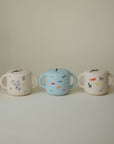 Three mushie Snack Cups with lids and handles are arranged in a row. Perfect for toddlers, they feature spill-proof designs: blue floral on the left, colorful animals in the center, and animal and nature prints on the right.