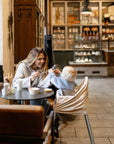 A woman smiles at a baby in a highchair, playfully holding out a spoon. A mushie Rocket Rattle Teether rests on the table beside coffee and pastries in the cozy café, with shelves of baked goods in the background.