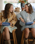 Two young girls in patterned jackets and dresses sit on wooden stools in a kitchen, smiling as they hold blue plates and the mushie Dinnerware Fork and Spoon Set. One wears a hair bow, both enjoying their non-toxic plastic utensils.