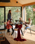 A woman serves food to three children using the mushie Dinnerware Fork and Spoon Set in a modern, sunlit room with large windows. A chair is decorated with a red bow, and trees are visible outside.