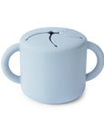 The mushie Snack Cup in Powder Blue is a spill-proof cup with two handles and a flexible, slit lid that lets little hands reach snacks while preventing spills.