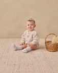 Baby in Rylee + Cru Bubble Knit Cardigan sits on a rug by a straw-filled wicker basket.