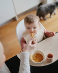 A baby in a high chair wearing a bib is being fed by an adult. The tray holds a bowl, cup, and is protected by a mushie Silicone Placemat. In the background, a cat walks near the window.