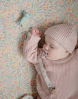 A baby in a pink knit sweater and hat sleeps in a crib with a floral-patterned sheet, holding a pacifier clip, with the mushie Dog Figurine Teether nearby.