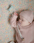 A baby in a pink knit sweater and hat sleeps in a crib with a floral-patterned sheet, holding a pacifier clip, with the mushie Dog Figurine Teether nearby.