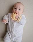 A baby in a white onesie lies on a beige surface, holding and chewing on the mushie No-Drop Sun Teether (Sunshine).