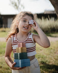 Smiling girl outdoors holds mushie Nesting Stars Toy, stacking a star to her eye.