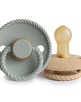 The Mushie FRIGG Rope Natural Rubber Pacifier 2-Pack includes muted green and beige pacifiers with round shields, ring handles, and rope-like textured edges. The beige pacifier is pictured lying on its side.