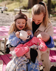 A toddler smiles outside, holding a mushie Snack Cup while a woman holds a baby.