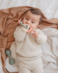A baby in a cream knit outfit lies on beige and tan blankets, holding and chewing on the mushie Dino Teething Ring. A green sensory toy rattle is nearby.