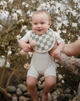 A smiling baby enjoys a joyful moment outdoors, held by tattooed adult arms in front of blooming branches, while playing with the mushie Silicone Baby Rattle Toy.