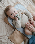 A baby in a beige outfit lies on a padded mat, smiling and chewing on the green mushie Dog Figurine Teether, while an adult’s hands gently rest on the baby’s belly.