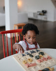 A young child practices fine motor skills while focusing on placing colorful letters into the Mushie Wooden Puzzle in a bright, modern living room.