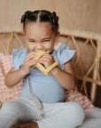 A young child with braided hair sits on a wicker chair, smiling and playfully biting the mushie No-Drop Sun Teether (Sunshine) while wearing a light blue shirt and white pants.