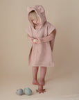 A young child wears the mushie Bear Poncho Towel in soft pink, barefoot and looking down while holding a toy. Three animal-shaped toys are scattered on the beige floor near their feet.