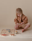 A young child with blonde hair sits on the floor in neutral-toned clothes, playing with the Mushie Wooden Puzzle. The educational toy helps develop fine motor skills, set against a calm beige background for a minimalistic look.