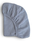 The mushie Extra Soft Bassinet Muslin Crib Sheet, shown folded in light gray with visible elastic edges, features a soft, crinkled muslin texture—ideal for breathable and cozy baby bedding.