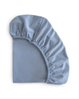 The mushie Stretchy MINI Crib Sheet in light blue is displayed with one corner turned up, revealing its elastic edge and soft organic cotton fabric on a white background.