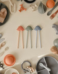 Mushie Training Chopsticks are surrounded by assorted silicone baby utensils and toys.