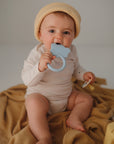 Baby in beige outfit chews on Mushie Animal Ring Teether while sitting on blanket.