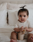 A baby with dark hair in a topknot sits on a white bed wearing a cream outfit, holding a soft toy and cup, with a FRIGG Natural Rubber 6-Pack of pacifiers by their side, looking at the camera.