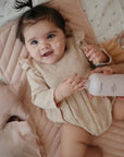 A smiling baby with a dab of cream on their cheek lies on a soft mat, holding a pink Mustela bottle as an adult applies cream. Nearby are the mushie Star Lovey Blanket and a plush star toy, creating a cozy scene.