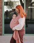 A woman with orange hair and glasses carries a baby in a pink wrap carrier. The baby holds a FRIGG Moon Natural Rubber Pacifier from FRIGG as they stand outside in front of large windows and look at the camera.