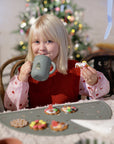 Child with Mushie Silicone Training Cup + Straw enjoys treats by Christmas tree.