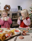 Two girls smile, sipping from Mushie Silicone Training Cups + Straws at a festive table.