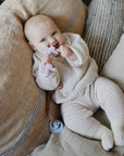 Happy baby in mushie knit outfit with Linen Pacifier Clip 2-Pack, holding flower teether.