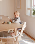 A young child with blond hair sits in a high chair at a wooden table, eating with a spoon. They wear the mushie Linen Pacifier Clip 2-Pack on their shirt in a cozy, sunlit room with a plant on the windowsill.