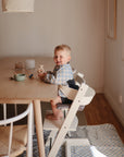 A young child sits in a white high chair at a wooden table, smiling with a toy. The Mushie Splat Mat protects the floor beneath, while cups and bowls are on the table in natural light from a nearby window.