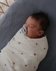 A baby sleeps in a mushie Organic Cotton Muslin Swaddle Blanket with a pacifier on a gray blanket.