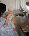In a modern kitchen, a woman in a blue checkered dress holds her smiling baby in a knit hat and outfit as they use the Mushie Silicone Fresh Food Feeder for self-feeding. A baby food tray rests on the counter nearby.