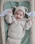 Smiling baby in a bunny-ear hat cuddles the mushie Figurine Teether on a soft blanket.