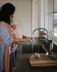 A woman in a blue checkered dress holds her baby in a mushie Baby Wrap while pouring water into the kitchen sink. Sunlight shines through the window, and baby items are scattered on the counter.