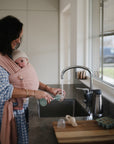 A woman in a blue checkered dress washes baby items at the kitchen sink using the mushie Silicone Baby Bottle Brush while carrying a baby in a pink wrap. The baby, wearing a white hat, looks at the camera as sunlight streams through the window.
