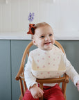 A smiling toddler in a red bow and cream bib with red designs sits in a wooden high chair, playing with her mushie Silicone Placemat made of food-grade silicone, while wearing a white long-sleeve shirt and red pants.