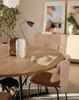 A cozy dining area features a round wooden table, beige chairs, and minimalist decor. A light pink shirt hangs over a chair, while a mushie Silicone Placemat adds charm to the setting as a small dog rests beneath the table.
