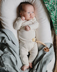 A baby in a cream outfit lies on a cushioned wicker bassinet, partially covered with the mushie Organic Cotton Muslin Swaddle Blanket in green floral; a wooden pacifier clip rests on the baby's waist.
