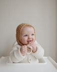 A smiling baby in a cream sweater and knitted bonnet sits in a high chair, holding the Mushie Silicone Fresh Food Feeder teething toy, against a neutral background.